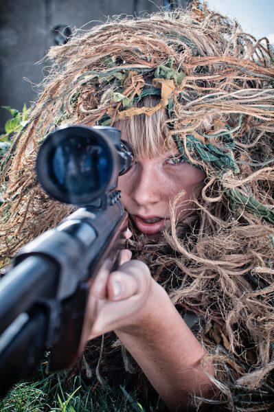 a person wearing a camouflage cover in his head and holding up a gun with scope