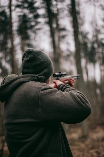 a hunter standing and aiming his gun by looking through the scope attached to his firearm